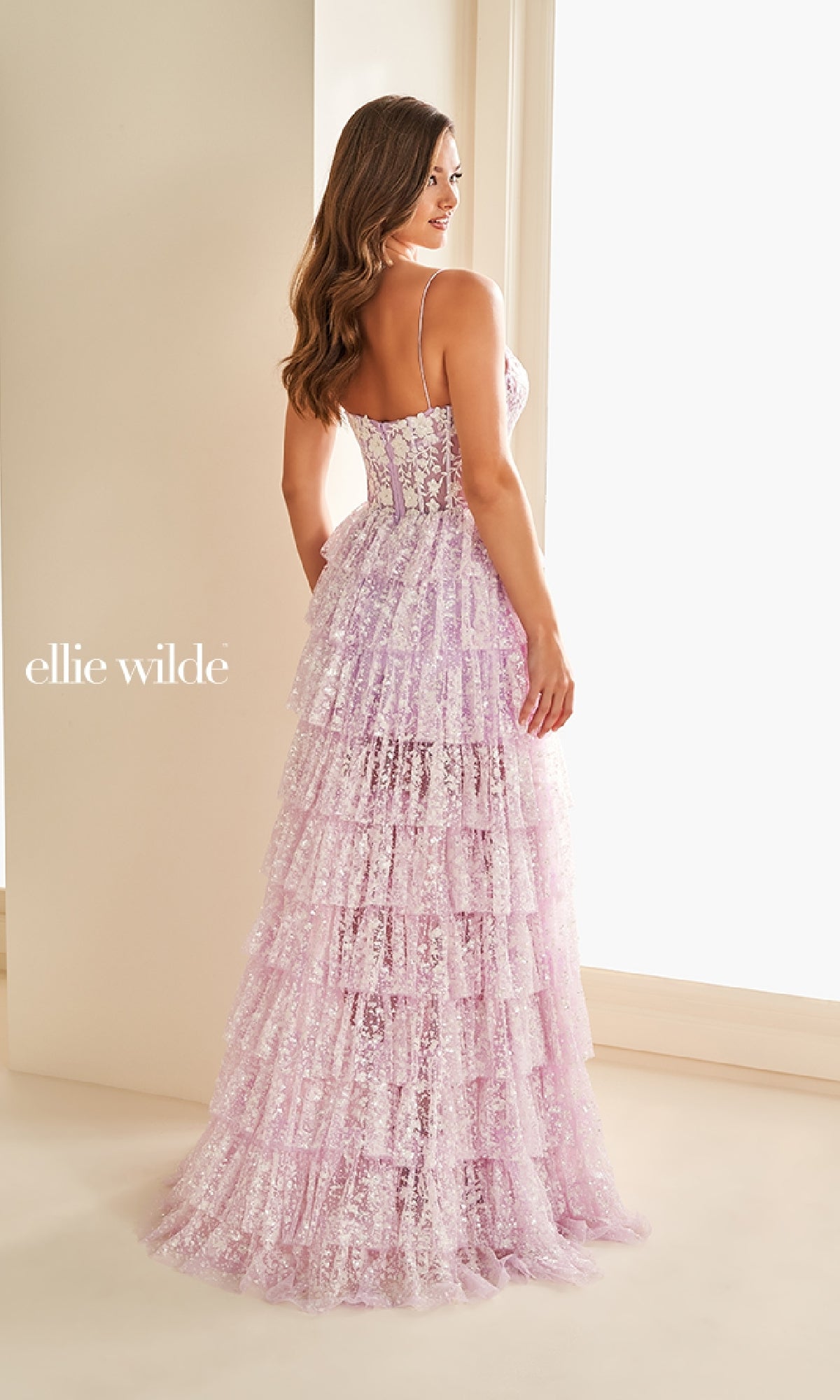 A woman stands indoors seen from the back, wearing the Ellie Wilde EW36216 long prom dress in lilac. The gown features spaghetti straps, a corset bodice, tiered ruffled layers, intricate lace appliqué, and exquisite craftsmanship.